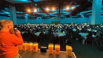 A Dynamic Events ireland host addresses a room filled with 750 Hexagon sales professionals at the Convention Centre Dublin, each holding up a glowing SolarBuddy light during the event finale. Yellow SolarBuddy collection bins are visible on stage, with the room lit by a soft blue ambience, capturing a powerful moment of unity and impact.