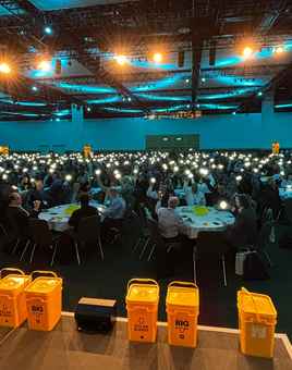 A Dynamic Events ireland host addresses a room filled with 750 Hexagon sales professionals at the Convention Centre Dublin, each holding up a glowing SolarBuddy light during the event finale. Yellow SolarBuddy collection bins are visible on stage, with the room lit by a soft blue ambience, capturing a powerful moment of unity and impact.